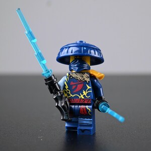 Peut inclure: Minifigurine Lego Ninjago bleue avec chapeau, masque et tenue bleus. La figurine tient une épée d'énergie bleue et une petite lame d'énergie. La tenue a des détails rouges et dorés.