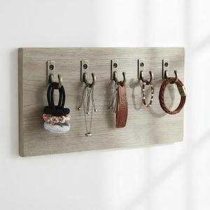 Könnte beinhalten: Eine rechteckige Wandhalterung in heller Holzoptik mit fünf Metallhaken in antikem Messington. An den Haken hängen verschiedene Accessoires, darunter Haargummis aus Stoff, eine silberfarbene Kette, ein braunes Lederband und verschiedene Armbänder.