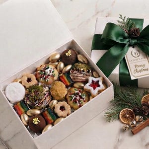 May include: A white gift box filled with assorted cookies, including star-shaped jam cookies, rainbow-striped bars, and chocolate truffles topped with gold leaf. A separate white box with a dark green ribbon and a Merry Christmas tag sits nearby.