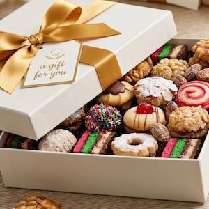 Pode incluir: Uma caixa de presente branca amarrada com uma fita dourada e uma etiqueta que diz "a gift for you". A caixa est&aacute; aberta, revelando uma variedade de biscoitos e doces coloridos, incluindo biscoitos arco-&iacute;ris e past&eacute;is cobertos com nozes.