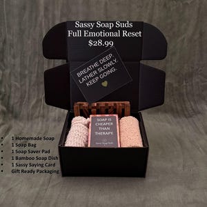 May include: A black gift box containing a wooden soap dish, a textured soap bag, a soap saver pad, and a bar of soap with a label reading SOAP IS CHEAPER THAN THERAPY. The box lid features the text Sassy Soap Suds Full Emotional Reset and a card reading BREATHE DEEP. LATHER SLOWLY. KEEP GOING.