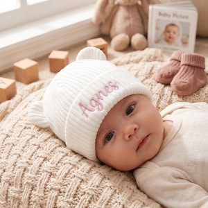 Puede incluir: Un gorro de bebé de punto acanalado blanco con pequeñas orejas de oso y el nombre Agnes bordado en hilo rosa en el puño delantero. El gorro está hecho de una tela suave y está diseñado para ajustarse cómodamente a la cabeza del bebé.