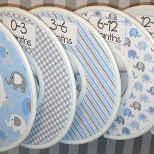 May include: Six blue and gray fabric-covered hangers with white trim and elephant print. The hangers are labeled with the following sizes: New Born, 0-3 months, 3-6 months, 6-12 months, 12-18 months, and 18-24 months.