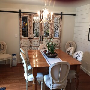 May include: A rustic dining room with a wooden table and six chairs. The chairs are upholstered in white fabric with a floral design. The table is set with a white runner and a centerpiece of white flowers in a brown bowl. The room has a white shiplap wall with framed botanical prints. A pair of distressed wooden doors with iron accents are visible in the background.