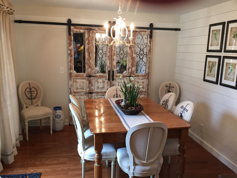 May include: A rustic dining room with a wooden table and six chairs. The chairs are upholstered in white fabric with a floral design. The table is set with a white runner and a centerpiece of white flowers in a brown bowl. The room has a white shiplap wall with framed botanical prints. A pair of distressed wooden doors with iron accents are visible in the background.