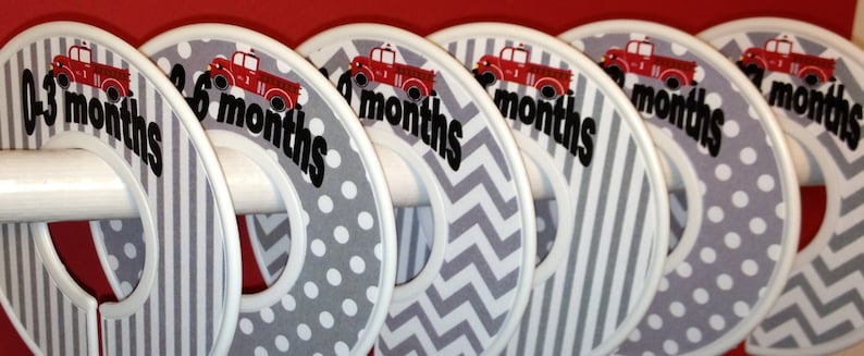 Puede incluir: Seis perchas de ropa para beb&eacute; de tela gris y blanca con dise&ntilde;os de camiones de bomberos rojos y texto negro que indica la edad en meses: 0-3 meses, 3-6 meses, 6-9 meses, 9-12 meses, 12-18 meses y 18-24 meses.
