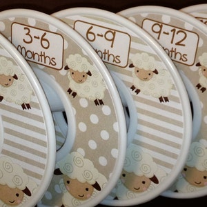 Puede incluir: Divisores de armario para ropa de bebé con diseño de oveja. Cada divisor está etiquetado con un rango de edad diferente en meses, de 0 a 3 meses a 18 a 24 meses. Los divisores tienen una combinación de colores beige y blanco con un patrón de rayas y lunares.