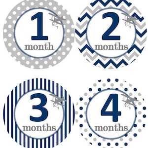 May include: Four round baby milestone stickers with different patterns. Each sticker features a navy blue number, a small airplane graphic, and the text "month" or "months". The patterns include gray and navy blue polka dots, gray and navy blue chevron, navy blue and white stripes, and gray and navy blue polka dots.