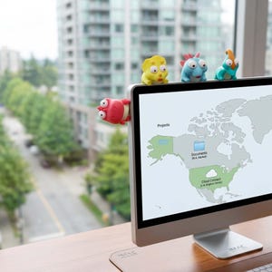 Könnte beinhalten: Ein Computermonitor mit einer Weltkarte auf dem Bildschirm. Mehrere bunte, cartoonartige Monster-Clips in Rot, Gelb, Blau und Türkis sind oben und an der Seite des Monitors befestigt.