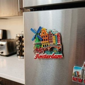 Könnte beinhalten: Ein farbenfroher Amsterdam-Kühlschrankmagnet mit einer Windmühle, Gebäuden, einer Brücke und einem Fahrrad. Das Wort "Amsterdam" ist rot geschrieben. Der Magnet ist an einem Kühlschrank aus Edelstahl befestigt.