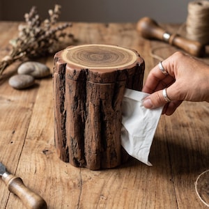 May include: A brown wooden tissue box shaped like a tree stump, with visible wood grain and bark texture. A hand is pulling a white tissue from the opening. The box sits on a wooden surface, suggesting a rustic or natural theme.