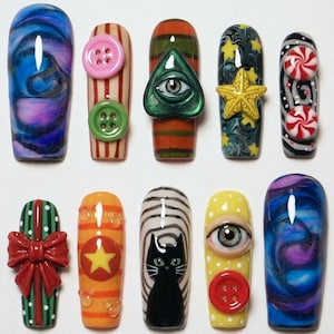 May include: A set of ten press-on nails featuring diverse designs including a blue and purple marble pattern, striped nails with buttons, a green eye charm, a yellow star, red and white peppermint candies, a red bow, and a black cat illustration.