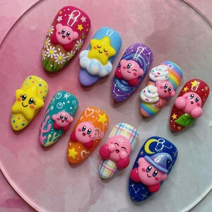 May include: A set of ten press-on nails featuring 3D character designs of a pink round creature. The nails display various colorful backgrounds including flowers, stars, rainbows, plaid patterns, and space themes with glitter and painted details.