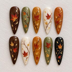 May include: A set of ten almond-shaped press-on nails featuring autumn-themed designs. Each nail displays painted maple leaves in shades of red and orange, delicate black branches, and scattered gold foil flakes over solid base colors of brown, olive green, cream, orange, and black.