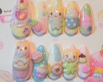 May include: A set of ten almond-shaped press-on nails featuring pastel colors and 3D charms. Designs include a sundae, strawberry, cat figures, a polka-dot mug, cupcakes, a donut, and a heart. The nails have glossy finishes with stripes, dots, and solid pastel backgrounds in shades of pink, blue, yellow, and green.