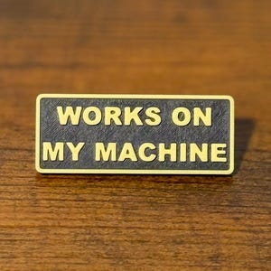 Könnte beinhalten: Ein rechteckiger Metallanstecker mit schwarzem, texturiertem Hintergrund und goldfarbenen, erhabenen Buchstaben, die WORKS ON MY MACHINE lesen. Der Anstecker hat einen dünnen goldenen Rand um den Text, der in zwei Zeilen angeordnet ist.