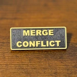 Könnte beinhalten: Eine rechteckige Anstecknadel mit einem dunkel strukturierten schwarzen Hintergrund und einem goldfarbenen Rand. Der Text MERGE CONFLICT ist in fetten, goldfarbenen Großbuchstaben in der Mitte der Anstecknadel zu sehen und klar lesbar.
