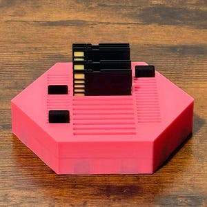 May include: A bright pink hexagonal storage stand featuring multiple slots for organizing small electronic memory cards. The stand has a ribbed surface texture with several black rectangular inserts and three black memory cards standing upright in the center.