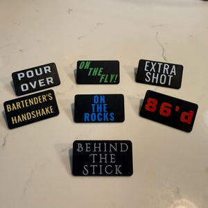 Puede incluir: Un conjunto de letreros rectangulares negros para bar, con texto blanco, verde, azul y rojo. Los letreros muestran frases como "POUR OVER", "BARTENDER'S HANDSHAKE", "ON THE FLY!" y "86'd".