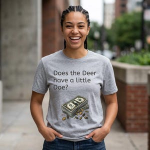 May include: A light gray t-shirt with the text "Does the Deer have a little Doe?" above a graphic of a stack of dollar bills and coins. The shirt is being worn by a person.