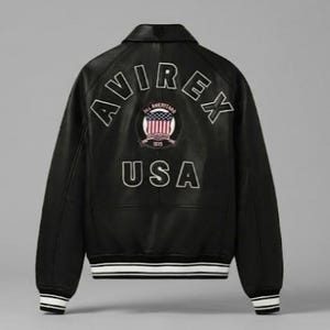 May include: A black leather varsity jacket featuring the text AVIREX in an arched white outline font across the upper back. Below is a circular patch with an American flag graphic and the text USA in large white outlined letters at the base.