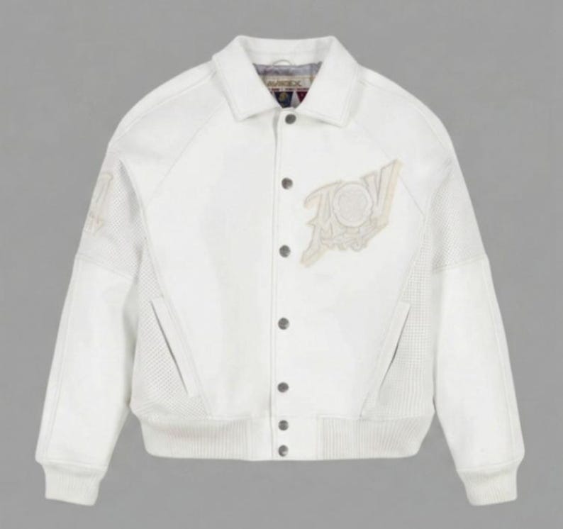 May include: A white varsity-style jacket featuring a classic collar, snap-button front closure, and ribbed cuffs and hem. The jacket includes a large embroidered patch on the left chest and textured side panels, with the Avirex brand label visible at the neck.