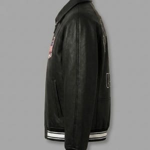 May include: A black leather varsity jacket featuring a classic collar, long sleeves, and a ribbed hem with white stripes. The jacket displays a rectangular patch on the chest and large embroidered lettering on the back, showcasing a structured fit.