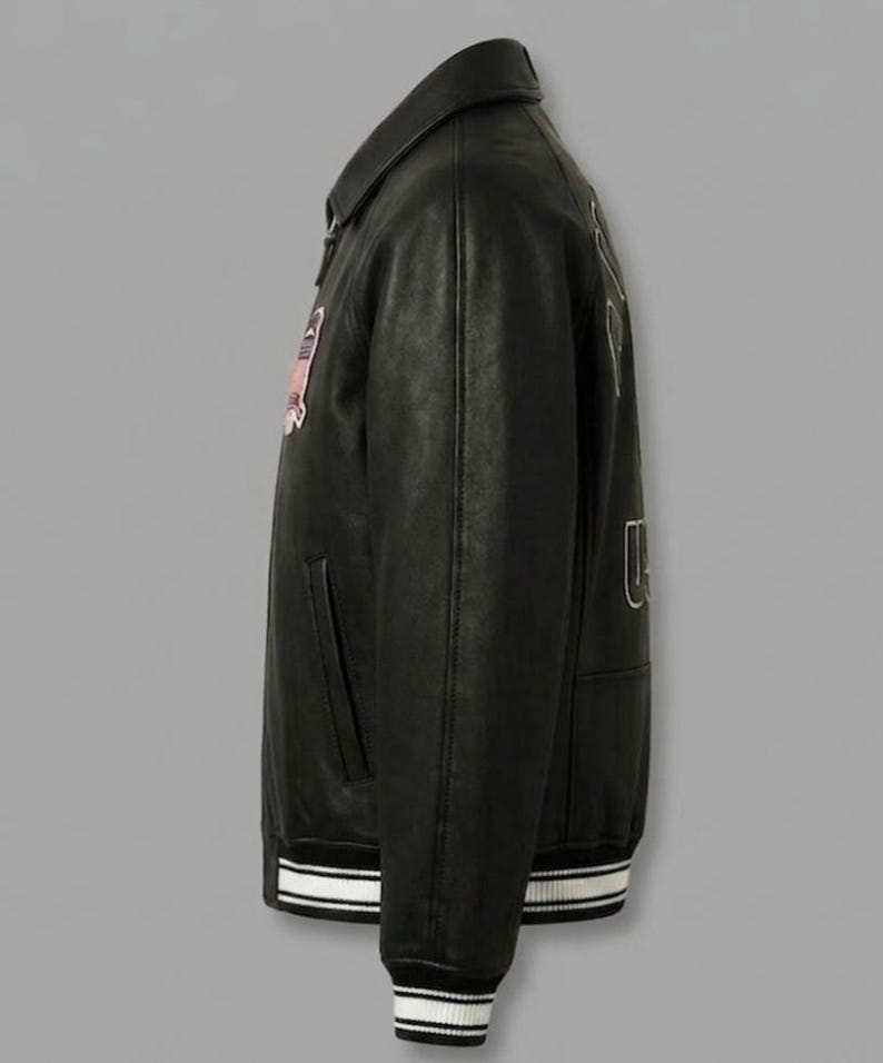 May include: A black leather varsity jacket featuring a classic collar, long sleeves, and a ribbed hem with white stripes. The jacket displays a rectangular patch on the chest and large embroidered lettering on the back, showcasing a structured fit.