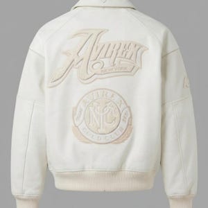 May include: An off-white bomber jacket featuring a large embroidered Avirex New York logo on the back. Below the text is a circular emblem with the letters NYC and the words Trade Avirex Gold Club. The jacket has a ribbed collar, cuffs, and hem.