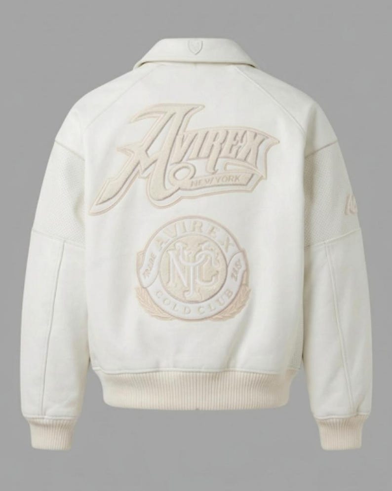 May include: An off-white bomber jacket featuring a large embroidered Avirex New York logo on the back. Below the text is a circular emblem with the letters NYC and the words Trade Avirex Gold Club. The jacket has a ribbed collar, cuffs, and hem.