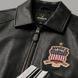 May include: A black leather jacket with a front zipper closure, a pointed collar, and white and black striped ribbed cuffs. An embroidered patch on the chest features the text AVIREX, a star, and a shield with stars and stripes, labeled EST. 1975.