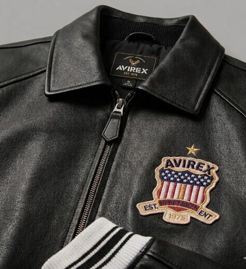 May include: A black leather jacket with a front zipper closure, a pointed collar, and white and black striped ribbed cuffs. An embroidered patch on the chest features the text AVIREX, a star, and a shield with stars and stripes, labeled EST. 1975.