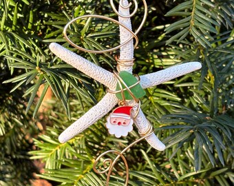 May include: A white starfish Christmas ornament with gold wire wrapped around it. A small green glass piece and a Santa Claus charm are attached to the wire. The ornament is hanging on a green pine tree.