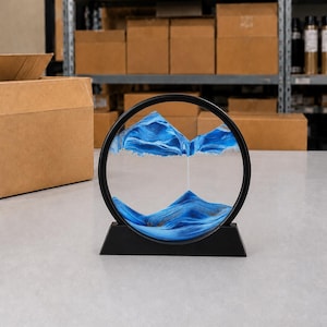 Puede incluir: Una pieza de arte de arena circular negra con arena azul que forma un paisaje montañoso. La arena está contenida dentro de un círculo de vidrio transparente, sostenido por una base negra. El fondo muestra cajas de cartón en estanterías.