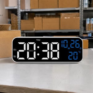 以下が含まれることがあります： 時刻20:38を表示する白いデジタル目覚まし時計。日付と温度も青色で表示されています。時計は角が丸い長方形で、黒い縁があります。背景には、段ボール箱のある倉庫の様子が写っています。