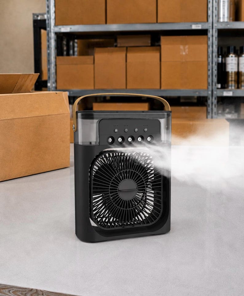 Puede incluir: Enfriador de aire port&aacute;til negro y gris con un asa marr&oacute;n. La parte delantera presenta un ventilador con un dise&ntilde;o circular. La parte superior tiene botones de control. Se emite niebla visible desde la parte delantera del enfriador. El fondo muestra estantes con cajas de cart&oacute;n.