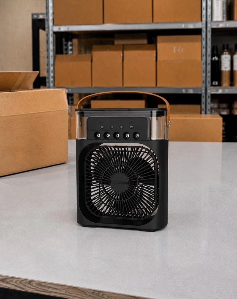 Puede incluir: Enfriador de aire port&aacute;til negro con una secci&oacute;n superior transparente y un asa marr&oacute;n. La parte delantera presenta un ventilador con un dise&ntilde;o circular. El enfriador se encuentra sobre una superficie blanca, con cajas de cart&oacute;n en el fondo.