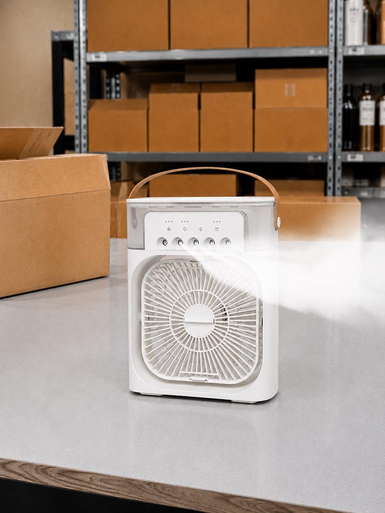 Puede incluir: Un enfriador de aire port&aacute;til blanco con un asa marr&oacute;n. La parte delantera presenta un ventilador con un dise&ntilde;o circular. El panel de control tiene varios botones e indicadores luminosos. Es visible una corriente de aire fresco, lo que sugiere que el dispositivo est&aacute; en funcionamiento.