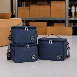 Puede incluir: Tres bolsas de almuerzo aisladas de color azul marino con asas negras y cremalleras plateadas. Cada bolsa tiene un logotipo circular y una correa para el hombro desmontable. Las bolsas son de diferentes tamaños y están dispuestas sobre una superficie gris.