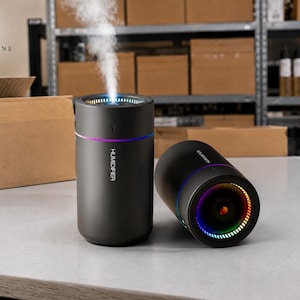 May include: Two black humidifiers with colorful LED lights. One is upright, emitting vapor, and the other is on its side. The word "HUMIDIFIER" is printed on the side of the upright humidifier. The background includes cardboard boxes and shelves.