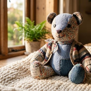 Może przedstawiać: Pluszowy miś typu patchwork wykonany z różnych tkanin, w tym niebieskiego dżinsu, kwiatowych nadruków i wzorów w kratę. Miś nosi pasujący szalik i ma małą prostokątną naszywkę na stopie z napisem EMILY'S BEAR 2024.