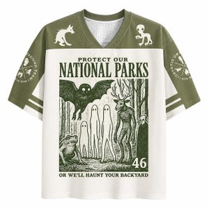 Könnte beinhalten: Ein olivgrünes und weißes V-Ausschnitt-Trikot mit dem Text PROTECT OUR NATIONAL PARKS OR WE'LL HAUNT YOUR BACKYARD. Die Grafik zeigt eine Fledermaus, einen Frosch, drei geisterhafte Figuren, ein Wesen mit Hirschkopf und eine vermummte Gestalt in einem Wald.