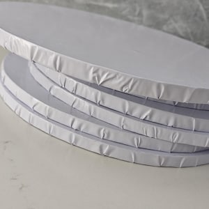 May include: A stack of four round white cake boards with smooth, flat surfaces and crimped, wrapped edges. Each circular board is constructed from a rigid material, featuring a clean, uniform white finish on the top and sides.