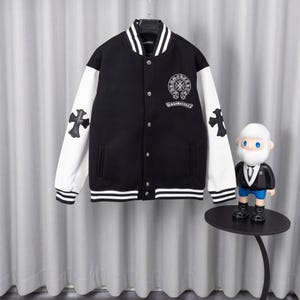 Pode incluir: Um casaco varsity preto e branco com corpo em lã preta, mangas brancas e acabamentos canelados às riscas na gola, punhos e bainha. O peito apresenta um logótipo circular bordado a branco com texto e cada manga tem um emblema de cruz preta.