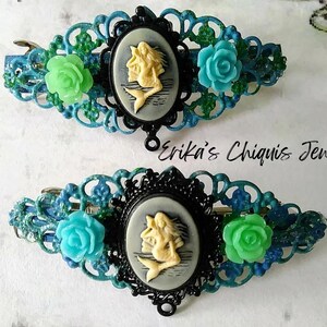 May include: Two blue and green hair clips with a cameo design featuring a mermaid. The cameo is set in a black filigree frame. The text "Erika's Chiquis Jewelry" is visible in the image.
