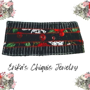 May include: A black and white striped cuff with red accents and red buttons. The cuff features a floral and skull pattern. The text "Erika's Chiquis Jewelry" is written in cursive below the cuff.