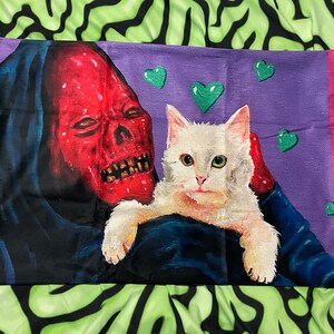 May include: A purple pillow with a red skull-faced figure holding a white cat. The background is decorated with green hearts.
