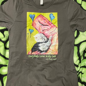 May include: A dark gray t-shirt with a graphic print of a pink and red creature holding a white cat. The graphic is surrounded by blue hearts and the text "Everybody Loves Kitty Cats" with the artist's name "art by Timothy Anglade".