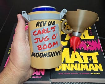 Puede incluir: Un enfriador de latas cilíndrico de color tostado con un borde superior gris y un accesorio en forma de embudo marrón con un detalle de llama roja. El frente muestra el texto: REV UP CARL'S JUG O' BOOM MOONSHINE en letras negras y rojas.