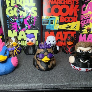 May include: A collection of seven stylized rubber ducks featuring various character designs, including one with a blue body and pink mohawk, a brown camo duck with purple goggles, and a duck wearing a black mask and heart-patterned base.